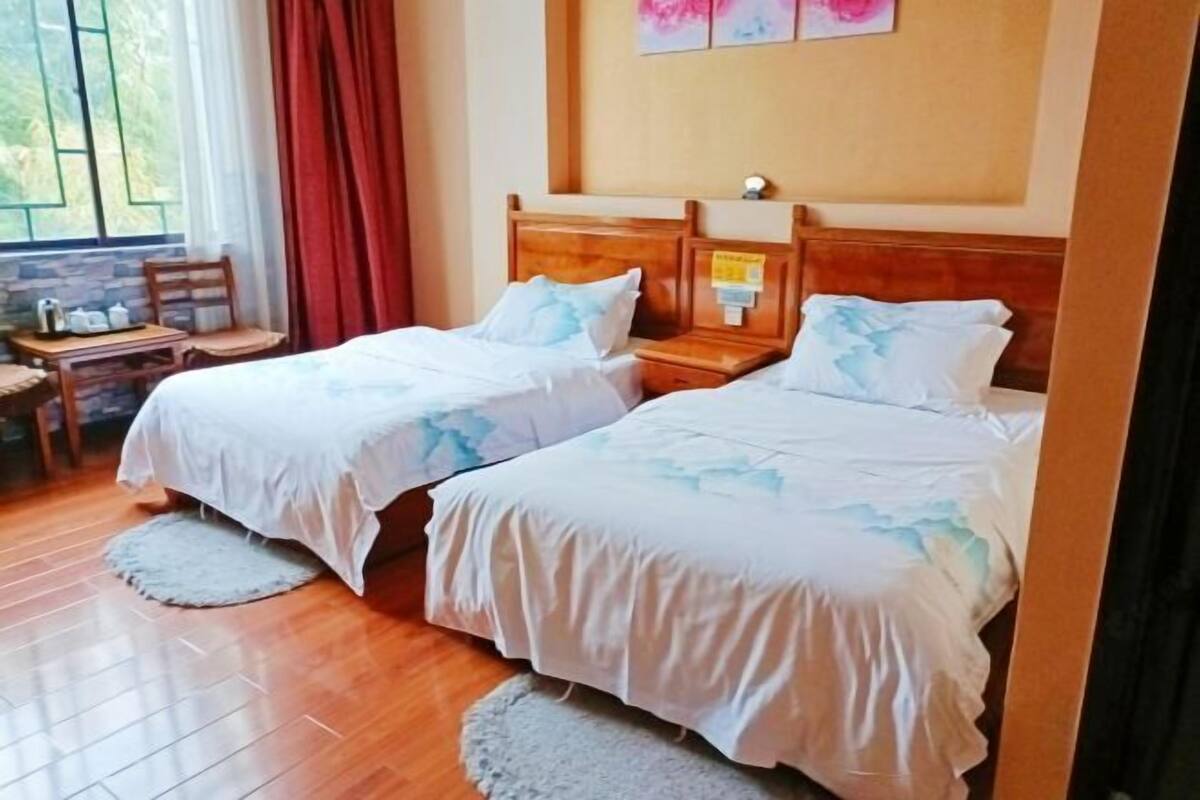 A room with two beds, a wooden headboard, a small table with a chair, and a window with curtains.