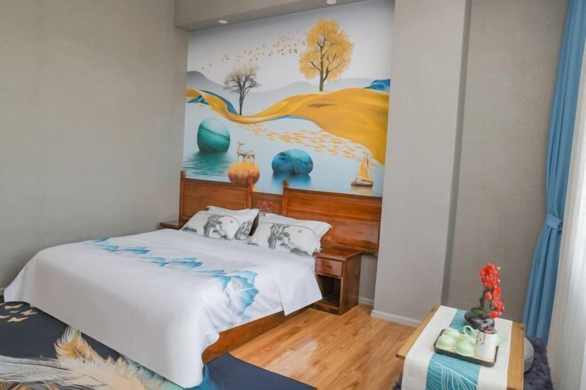 A bedroom with a bed, wooden headboard, a wall mural, a bedside table with a vase, and a window with curtains.
