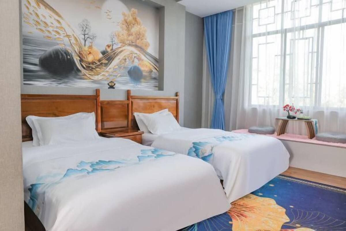 A hotel room with two beds, a wooden headboard, a large window with blue curtains, and a painting on the wall.