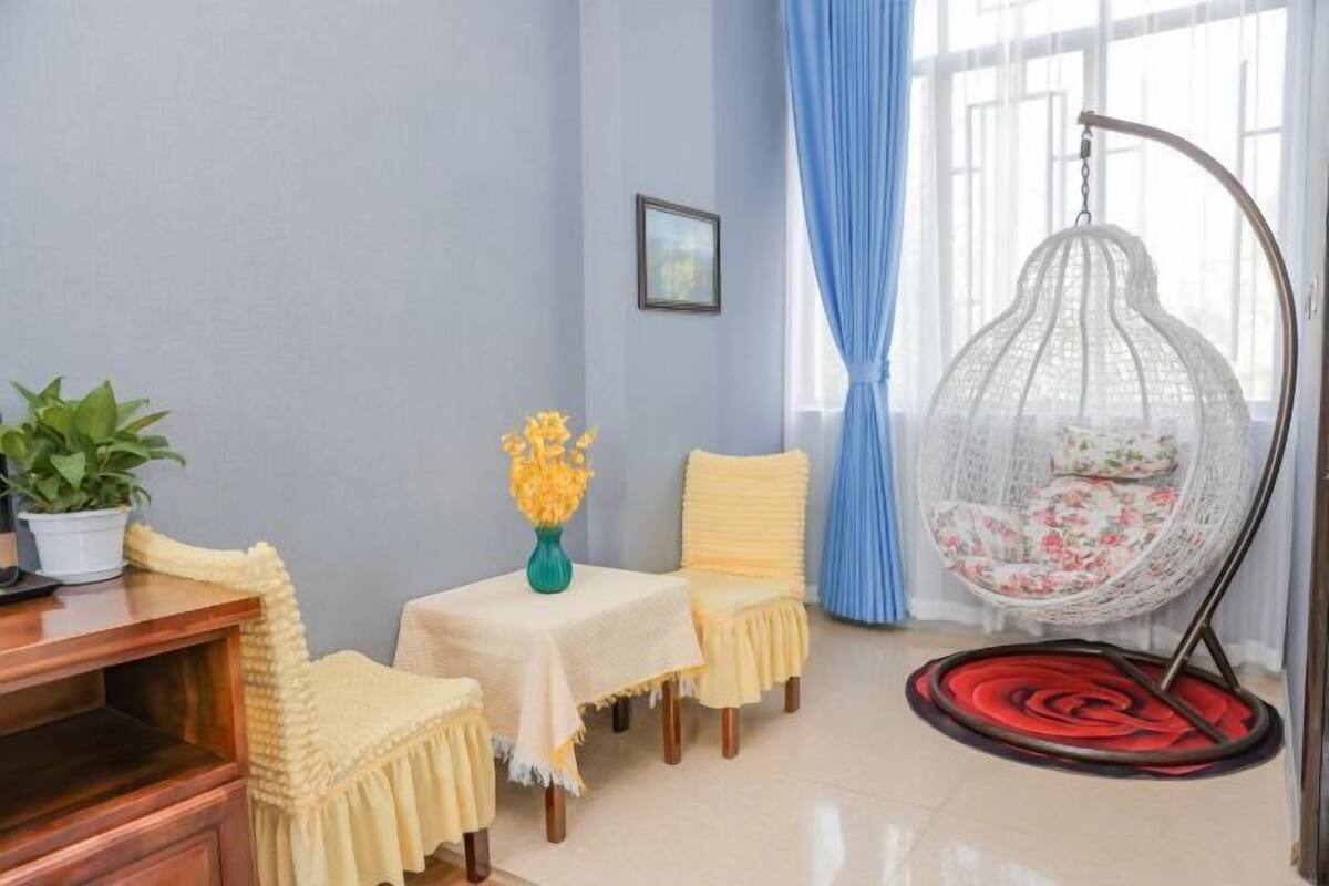 A room with a wooden desk, two chairs, a small table with a vase of yellow flowers, a hanging chair with a floral cushion, and a red and black rug.