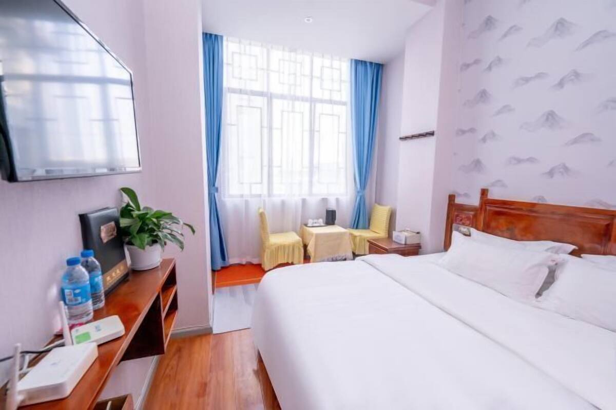 A hotel room with a bed, a desk with a computer, a chair, a small table, a plant, and a window with curtains.