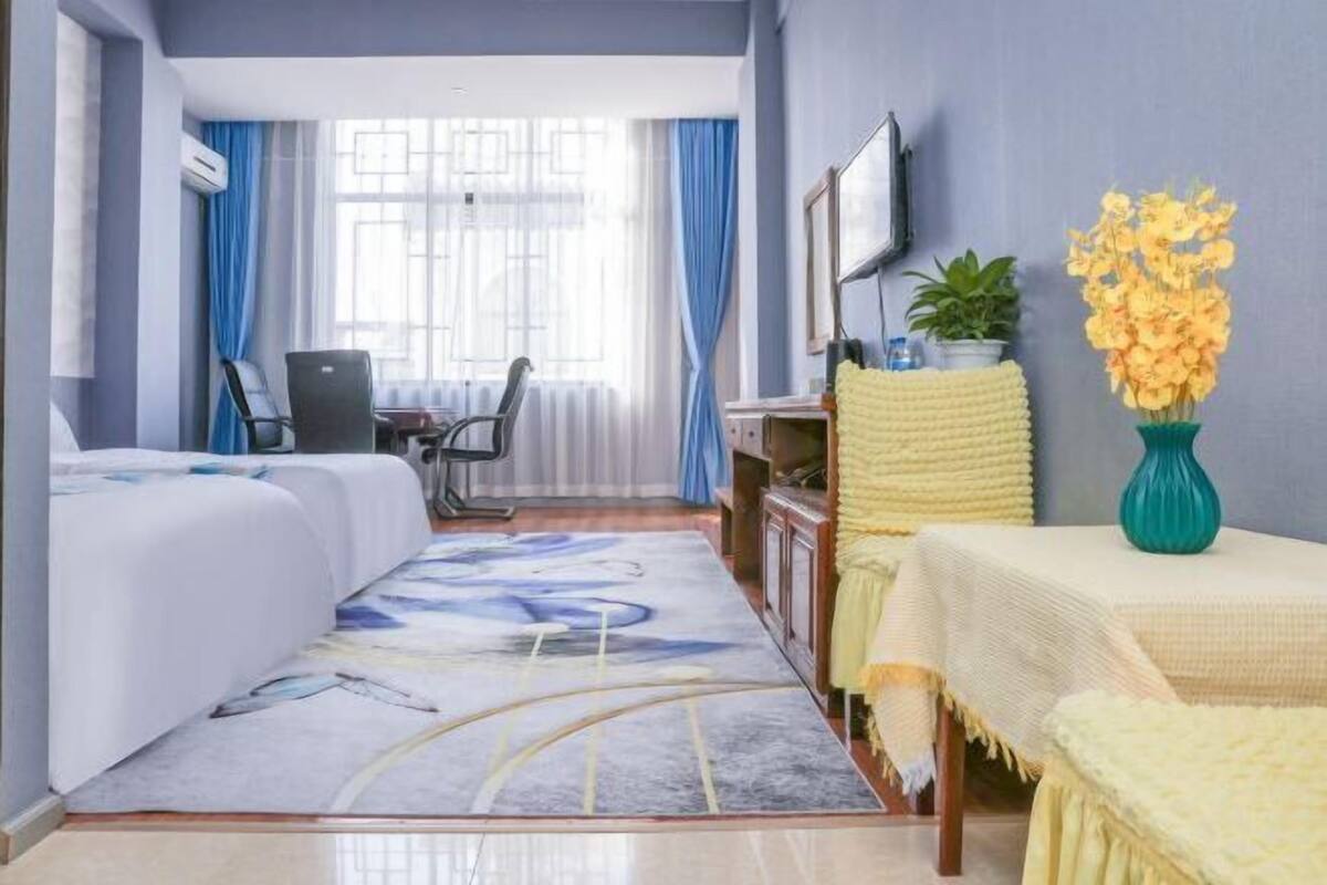 A hotel room with a bed, a desk, a chair, a TV, a window with curtains, and a vase with flowers.