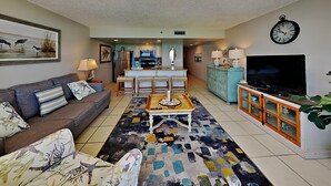 A living room with a sofa, coffee table, TV, and kitchen area in the background. - Edgewater Tower III #1109 by Resort Collection (Panama City Beach)
