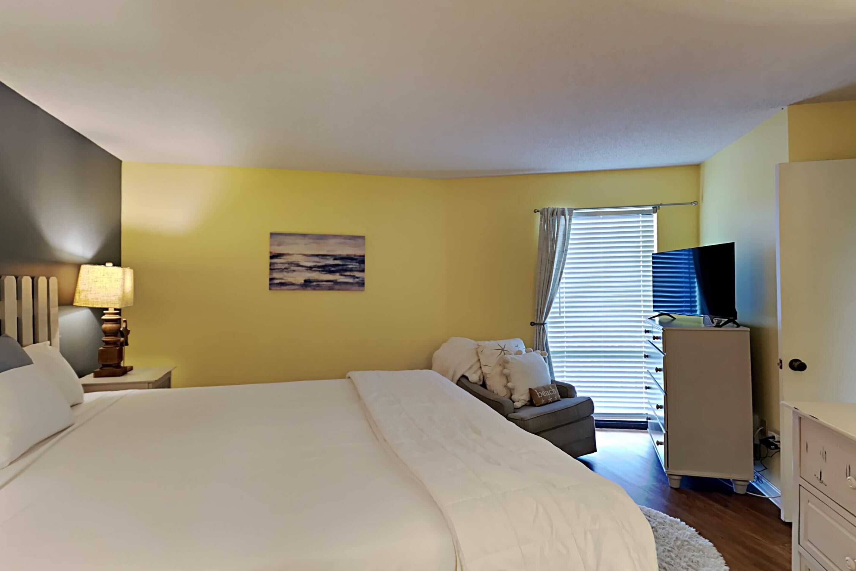 Une chambre à coucher comprenant un lit, une table de chevet, une lampe, une commode, un téléviseur et une fenêtre avec des stores.