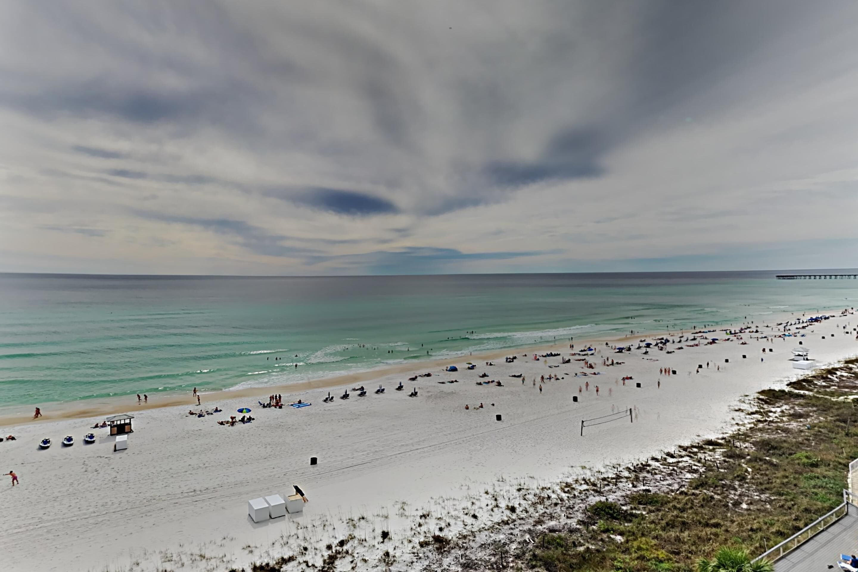 Une plage avec une longue jetée qui s’avance dans l’eau, une plage de sable où se trouvent des gens et des parasols, et un ciel nuageux.