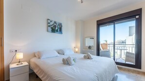 A hotel room with a large bed, a nightstand with a lamp, a painting on the wall, a balcony with a view, and a small table with chairs. - Modernly designed holiday flat in Mijas Costa (La Cala de Mijas)