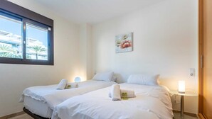 A hotel room with two beds, a window with a view of buildings, a painting on the wall, and a bedside table with a lamp. - Modernly designed holiday flat in Mijas Costa (La Cala de Mijas)