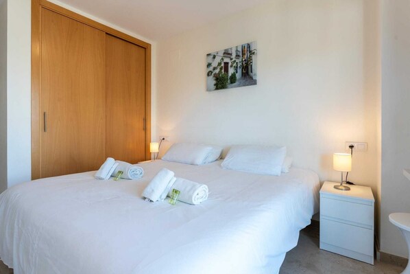 A neatly made bed with white linens and rolled towels, a bedside table with a lamp, and a wooden wardrobe.