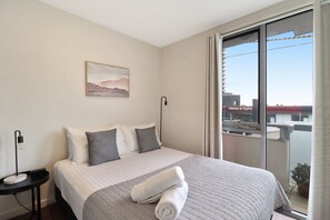 A hotel room with a bed, a nightstand, a painting, and a view of the city through the window.