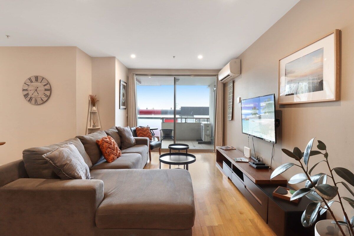 A modern living room with a sofa, a flat-screen TV, a coffee table, and a balcony with a view.