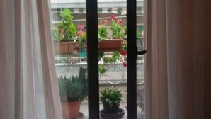 Een kamer met een bed, een raam met gordijnen en uitzicht op een balkon met plantenpotten.