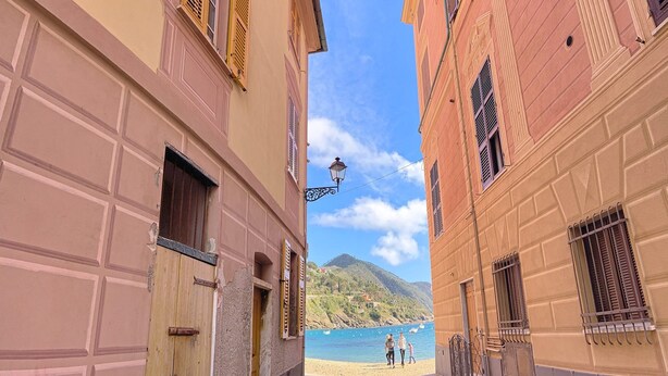 A narrow street with buildings on either side, a clear blue sky, and a distant view of a beach with boats.