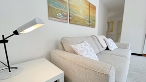 A living room with a beige sofa, a white side table, a floor lamp, and a painting of a boat on the wall.