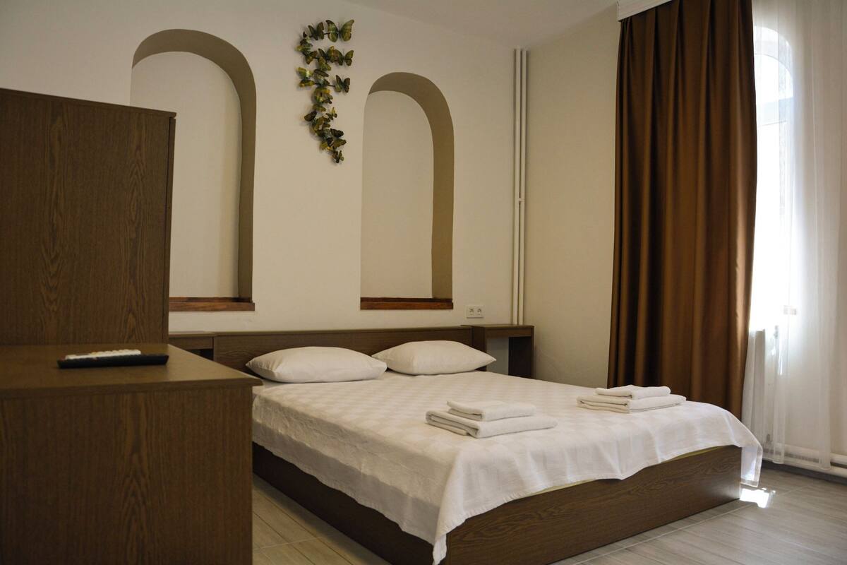 Ein Schlafzimmer mit einem Holzbett, weißer Bettwäsche, einer Fernbedienung auf einem Nachttisch aus Holz und einem Fenster mit braunen Vorhängen.