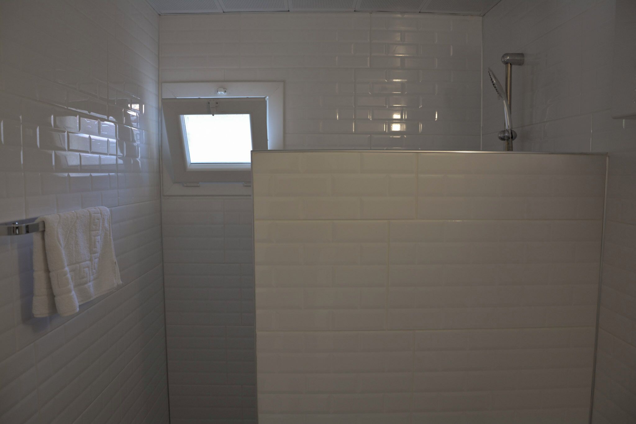 A bathroom with white tiles, a window, a shower, and a towel rack.