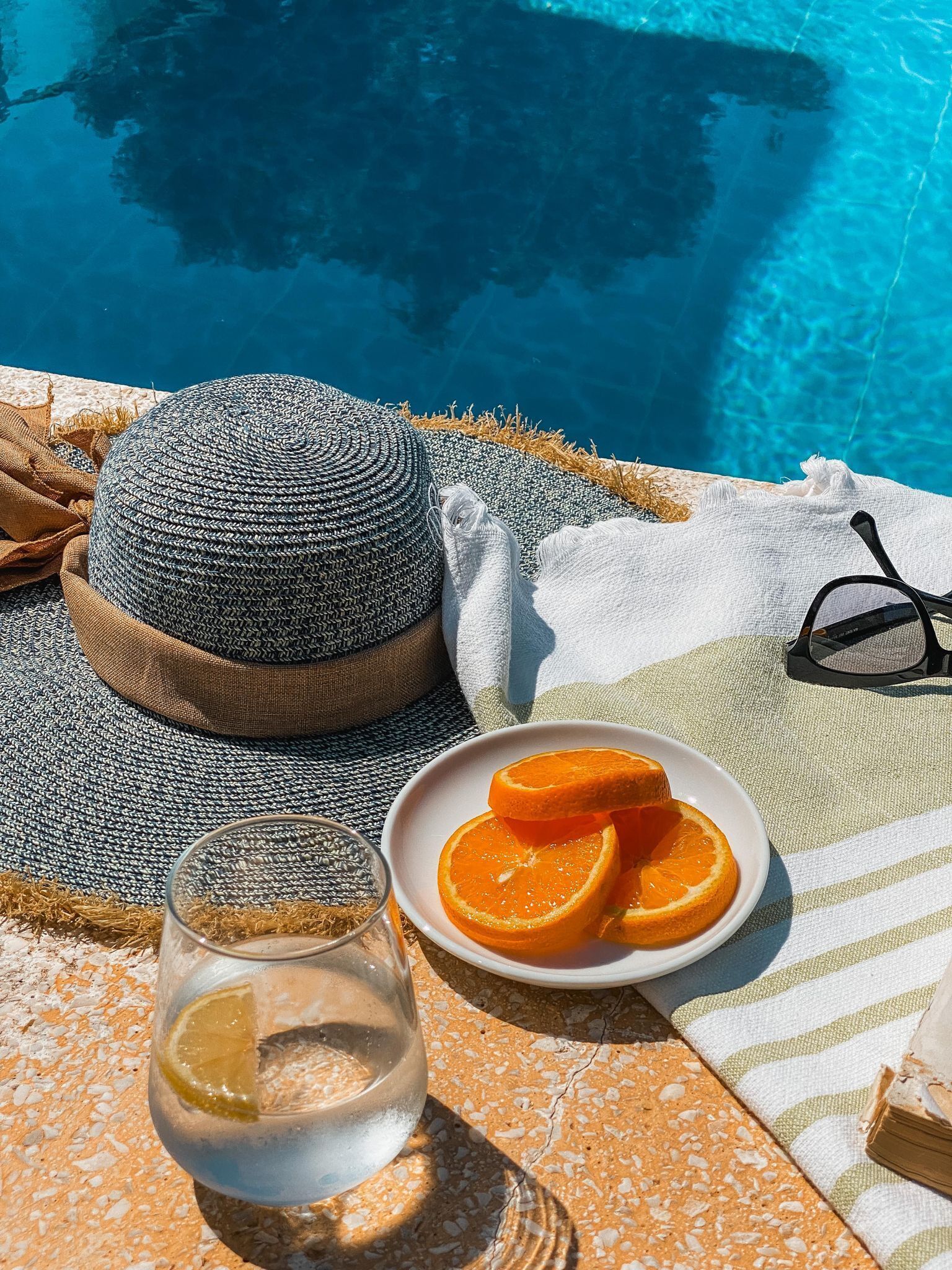 Cenário à beira da piscina com chapéu de palha, um prato de laranjas, um copo d'água e um par de óculos de sol.
