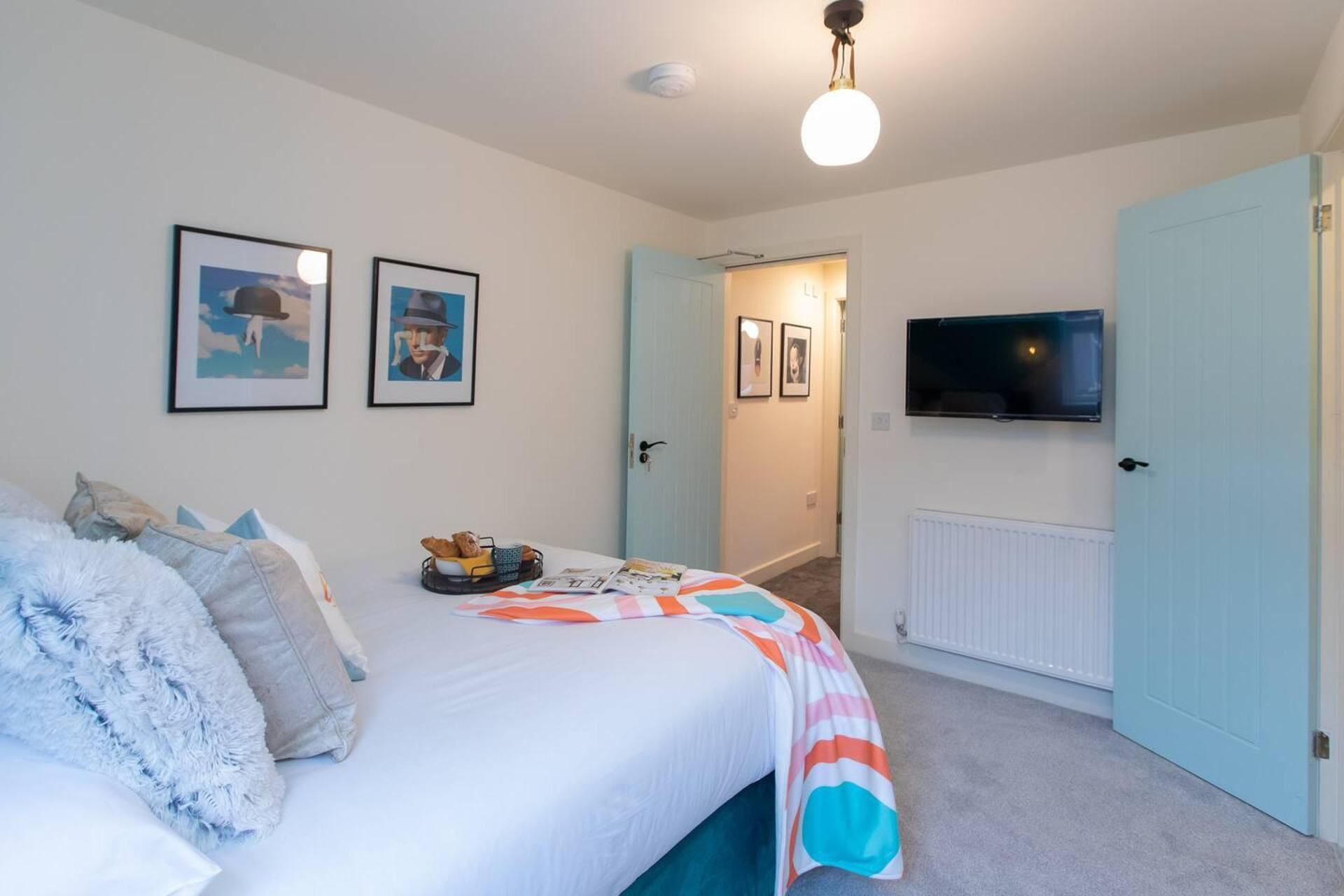 Une chambre à coucher avec un lit, des oreillers, une table de chevet sur laquelle se trouve un plateau de nourriture, un téléviseur fixé au mur et des tableaux encadrés accrochés au mur.