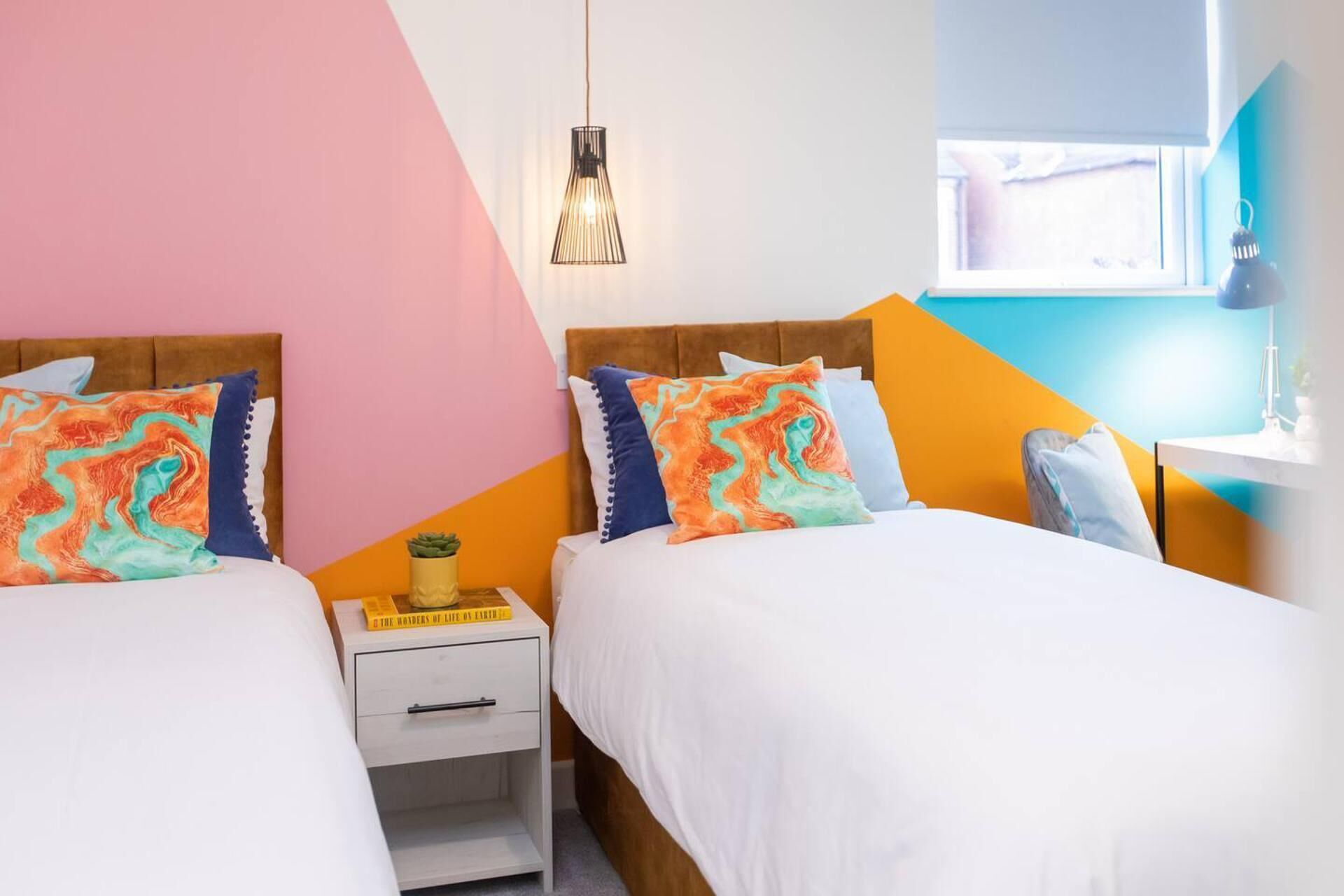 A bedroom with two beds, each with a white bedspread and colorful pillows. There is a white nightstand with a book and a small plant on it. A hanging light fixture is visible above the beds.