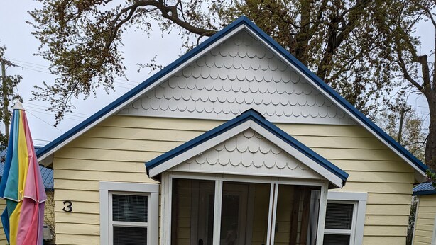 A small yellow house with a blue roof and white trim, featuring a porch with blue chairs and a table with a colorful umbrella. - Little Yellow Cottages #3 (Port Austin)
