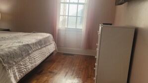 Un dormitorio con cama, mesita de noche, lámpara, ventana con persianas y refrigerador.