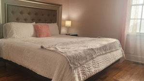 Una cama bien hecha con cabecera capitonada, una lámpara de noche y una ventana con persianas.