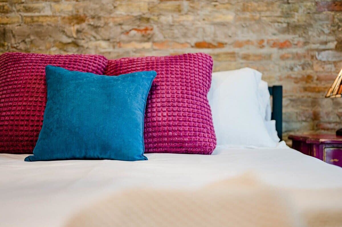 A bed with a white sheet and two pillows, one blue and one pink, against a brick wall.