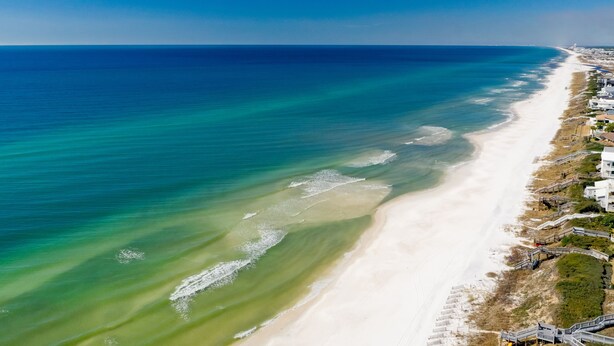 Eine Vogelperspektive zeigt einen Strand mit blauem Meer, einem sandigen Ufer und Gebäuden entlang der Küste.