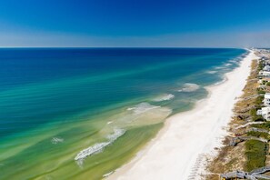 An aerial view of a beach with a clear blue ocean, a sandy shore, and buildings along the coastline.