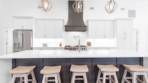Una cucina moderna con mobili bianchi, una grande isola con sgabelli, elettrodomestici in acciaio inossidabile e lampade a sospensione geometriche.
