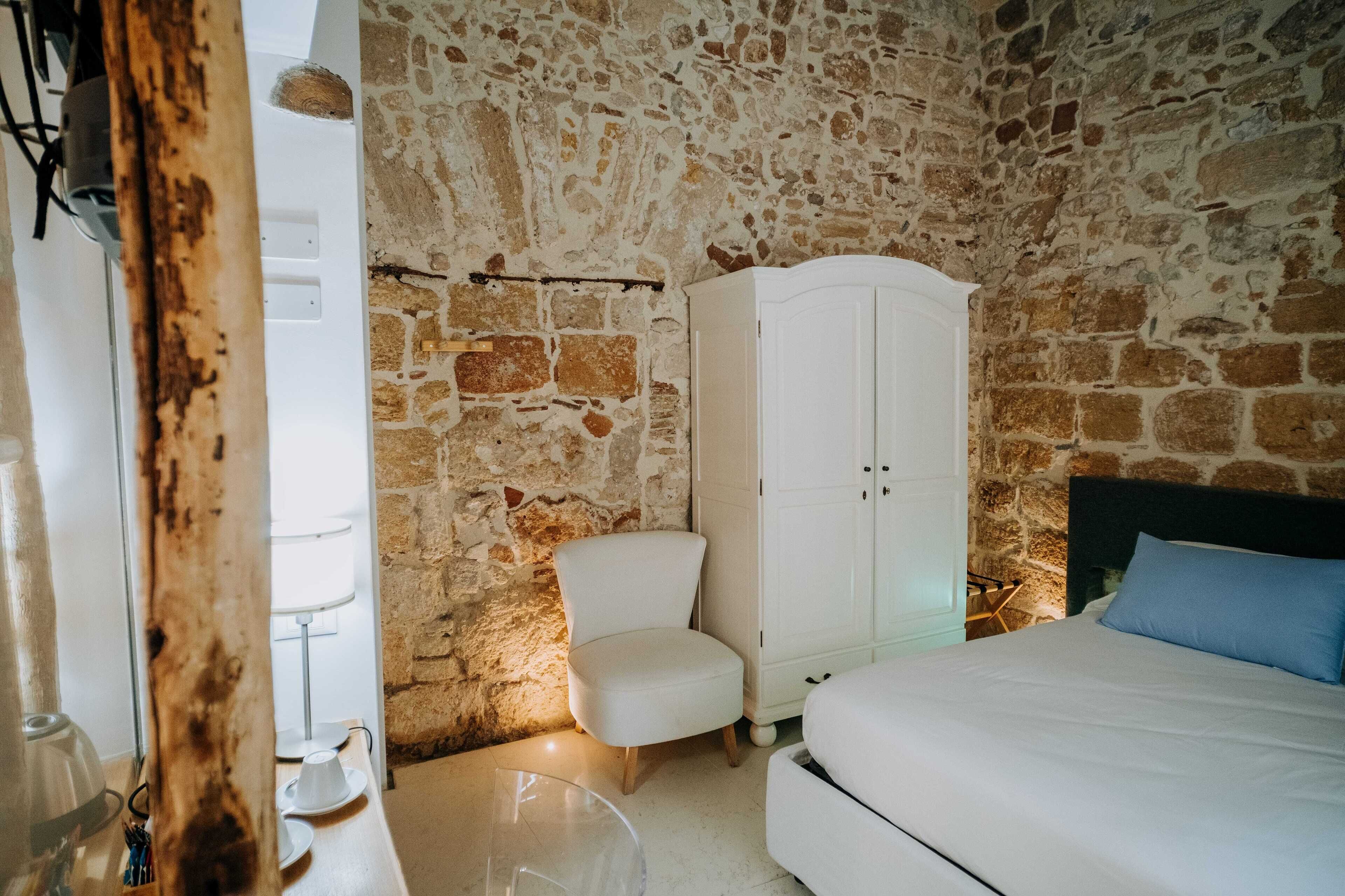 Un dormitorio con pared de piedra, un armario blanco, una silla blanca y una cama con almohada azul.