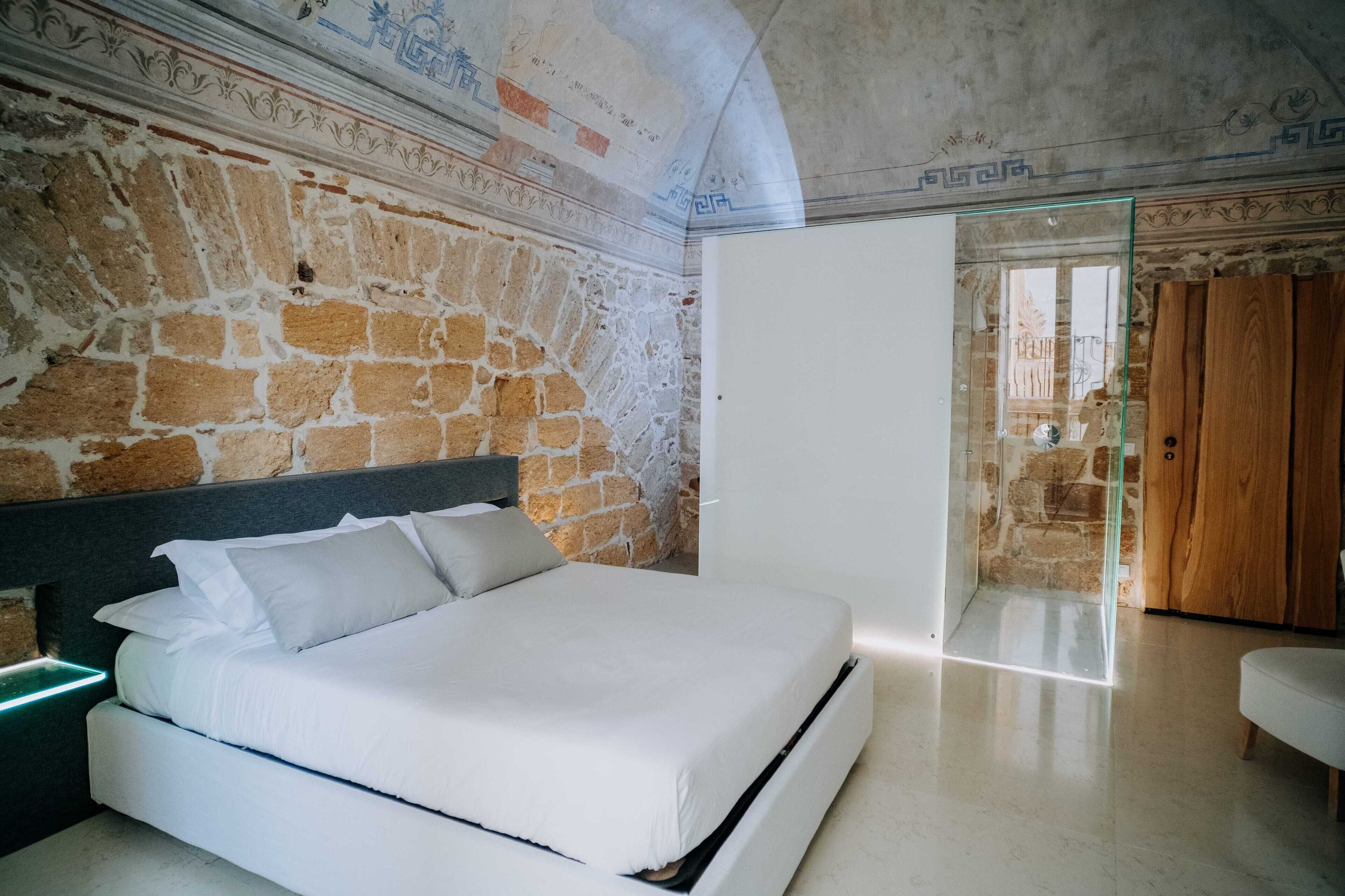 A modern bedroom with a stone wall, a bed with white bedding, and a glass door leading to a wooden door.