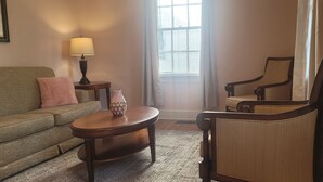 Uma sala de estar com sofá, mesa de centro, abajur, poltrona e janela com persianas.