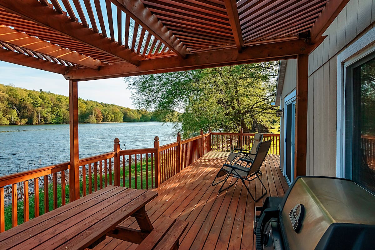 A wooden deck with a grill and chairs overlooking a lake.