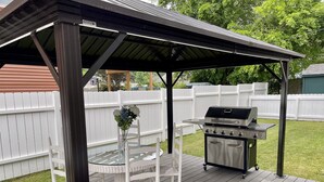 Un patio techado al aire libre con una parrilla, sillas de mimbre blanco y una mesa de vidrio.