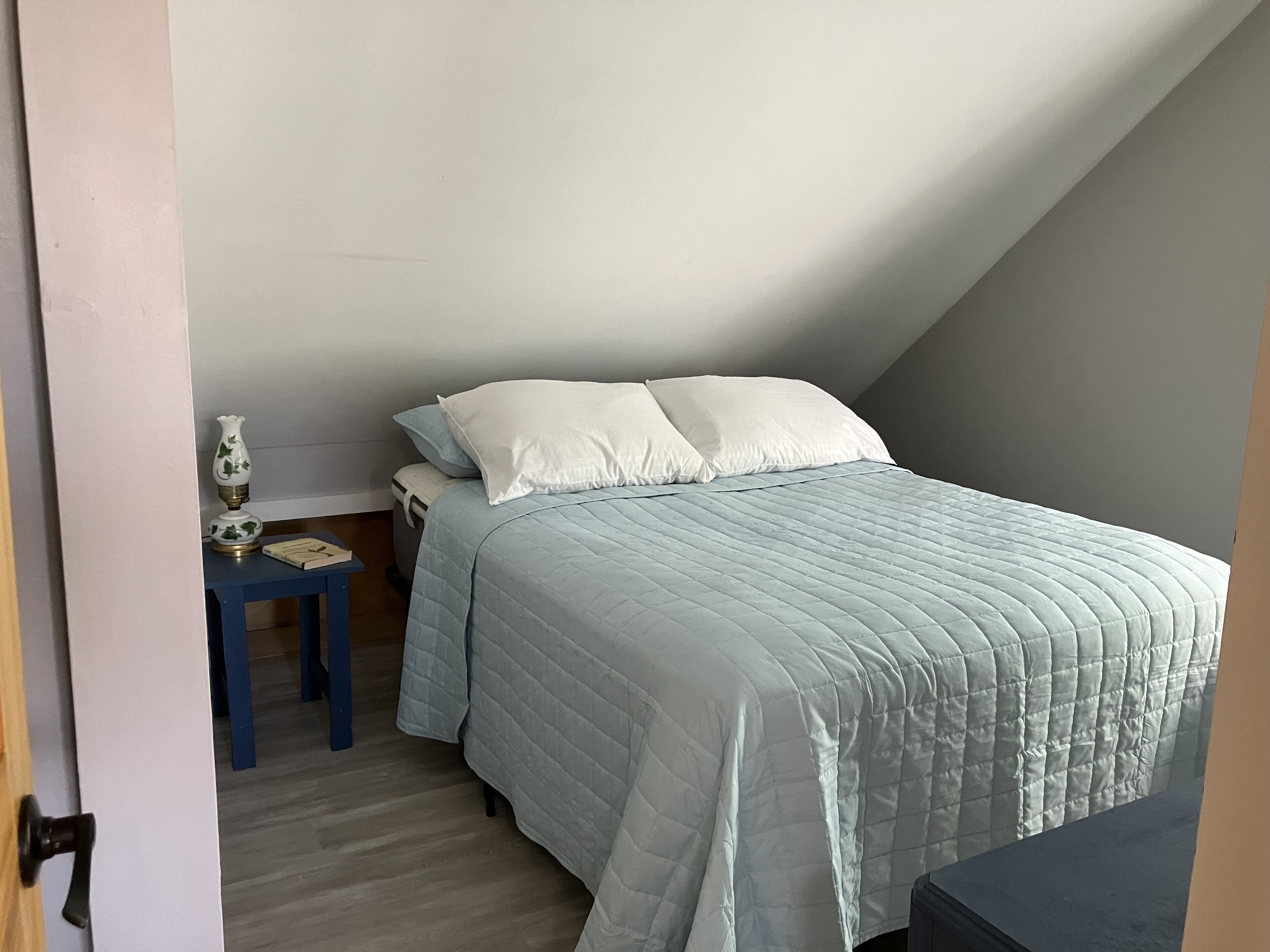 A small, neatly arranged bedroom with a bed, pillows, a nightstand, and a lamp.