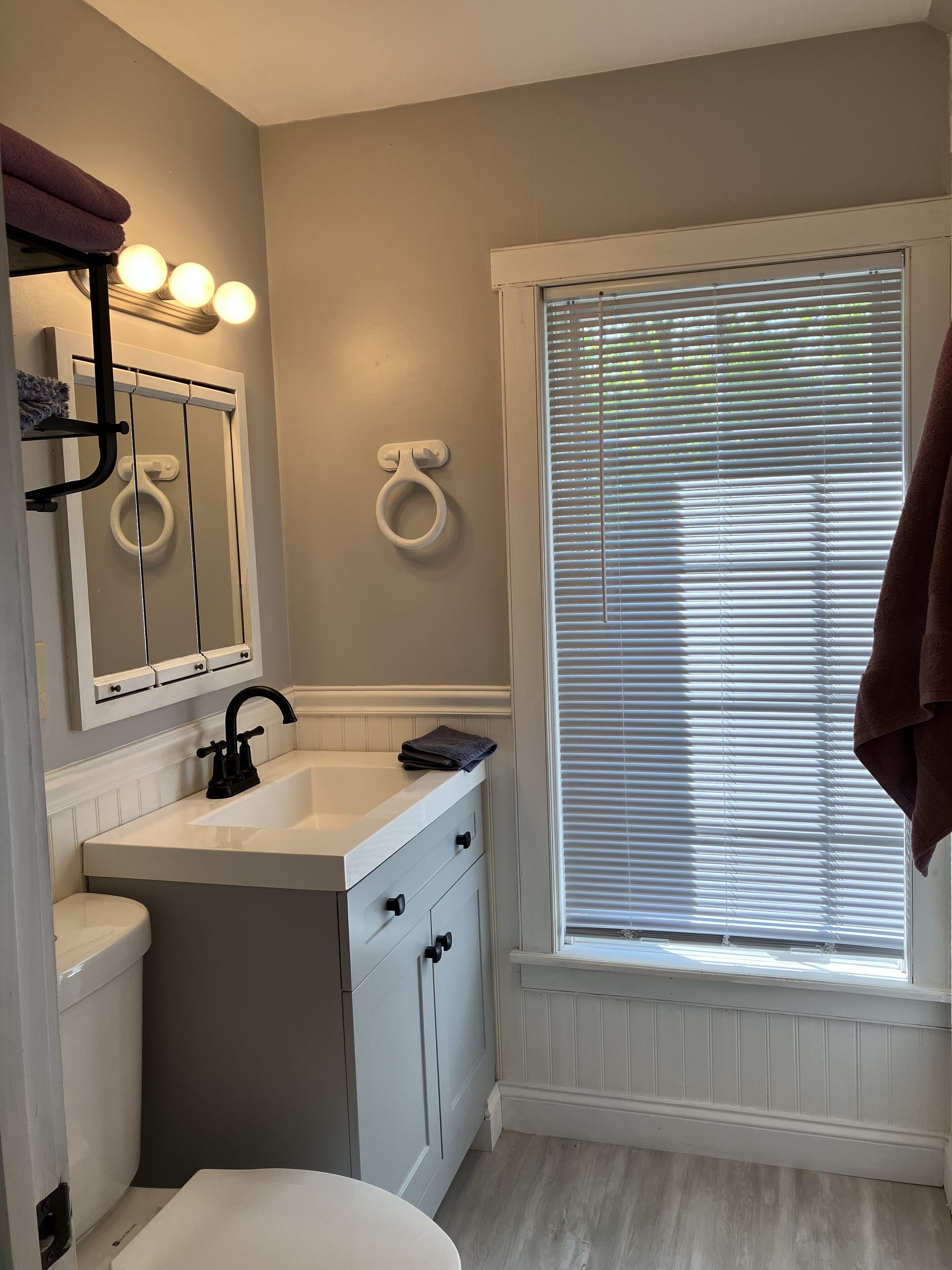 A bathroom with a white sink, vanity, and toilet, a window with blinds, and a towel rack with towels.