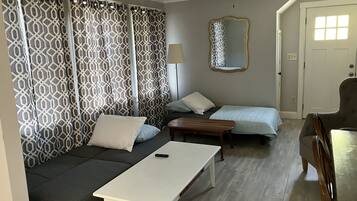 A compact room with a sofa, two single beds, a small table, a ceiling fan, and a mirror.