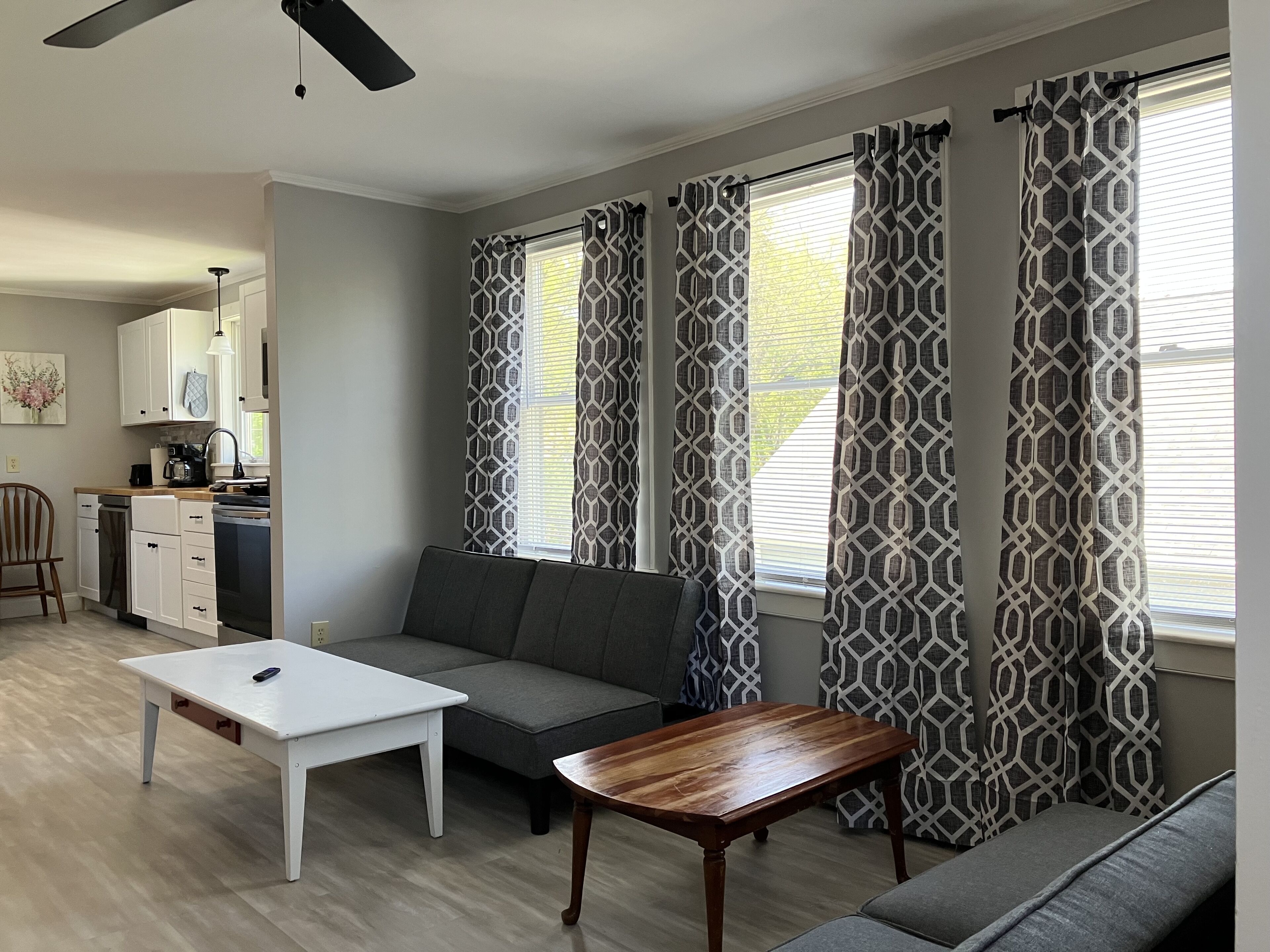 Ein modernes Wohnzimmer mit einem grauen Sofa, einem weißen Couchtisch und einem Holznachttisch. Es gibt eine Küchenzeile mit weißen Schränken und einer schwarzen Arbeitsplatte. Der Raum verfügt über ein gemustertes Vorhang und einen Deckenventilator.