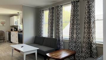 Ein modernes Wohnzimmer mit einem grauen Sofa, einem weißen Couchtisch und einem Holznachttisch. Es gibt eine Küchenzeile mit weißen Schränken und einer schwarzen Arbeitsplatte. Der Raum verfügt über ein gemustertes Vorhang und einen Deckenventilator.