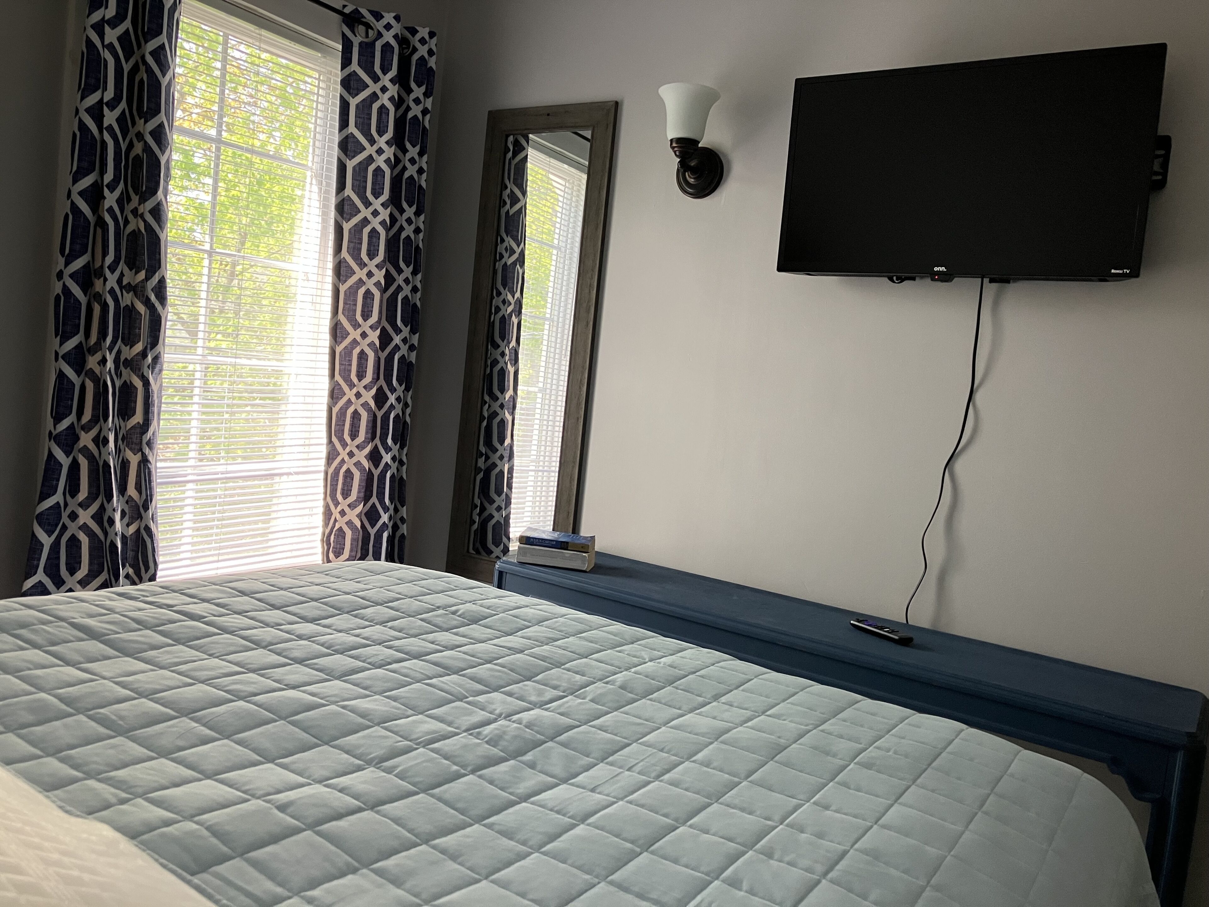 A bedroom with a bed, a TV mounted on the wall, and a window with patterned curtains.