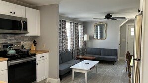 Eine moderne Küche mit Mikrowelle, Herd und Wasserkocher, ein graues Sofa, ein weißer Couchtisch und ein Deckenventilator.