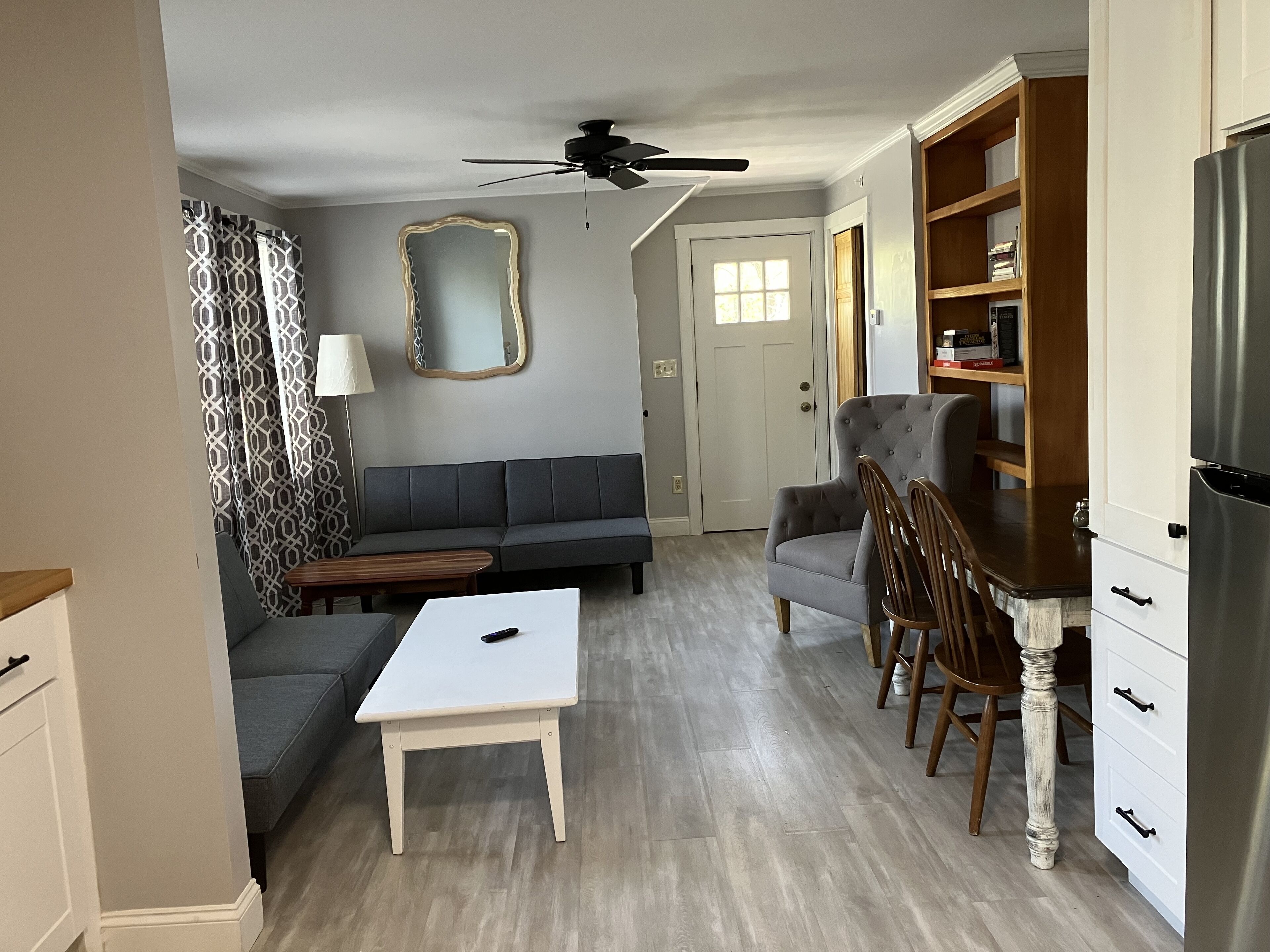 Un espacio habitable compacto con sofá, mesa de comedor, silla, ventilador de techo, refrigerador y estantería.