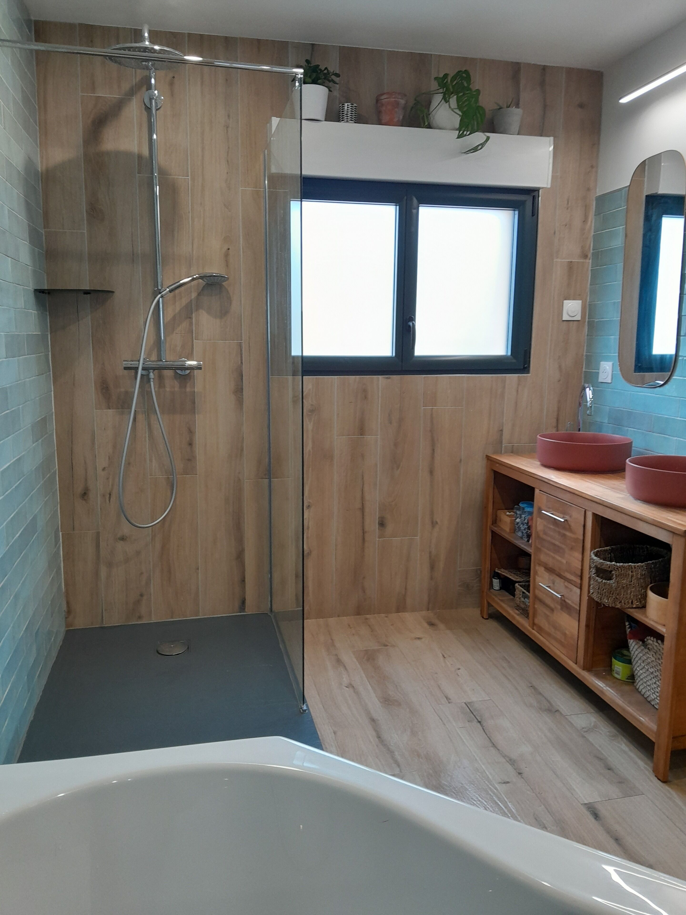 A modern bathroom with a glass-enclosed shower, wooden walls, a large window, and a wooden vanity with a red basin.