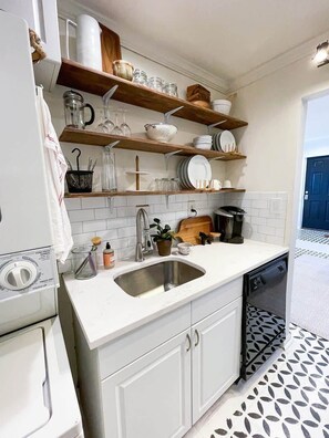A kitchen with white cabinets, a stainless steel sink, wooden shelves above the counter, and a tiled backsplash. - Charming Capitol Hill 1BR | Walk to Capitol+Patio (Washington)