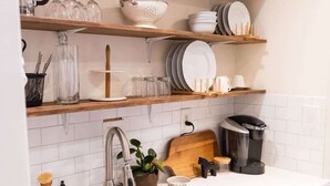 A kitchen with white cabinets, a stainless steel sink, wooden shelves with glassware, and a coffee maker.