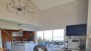En rummelig opholdsstue med stor sofa, sofabord og TV. Der er et moderne køkken, et spiseområde med udendørs siddepladser, og havudsigt.