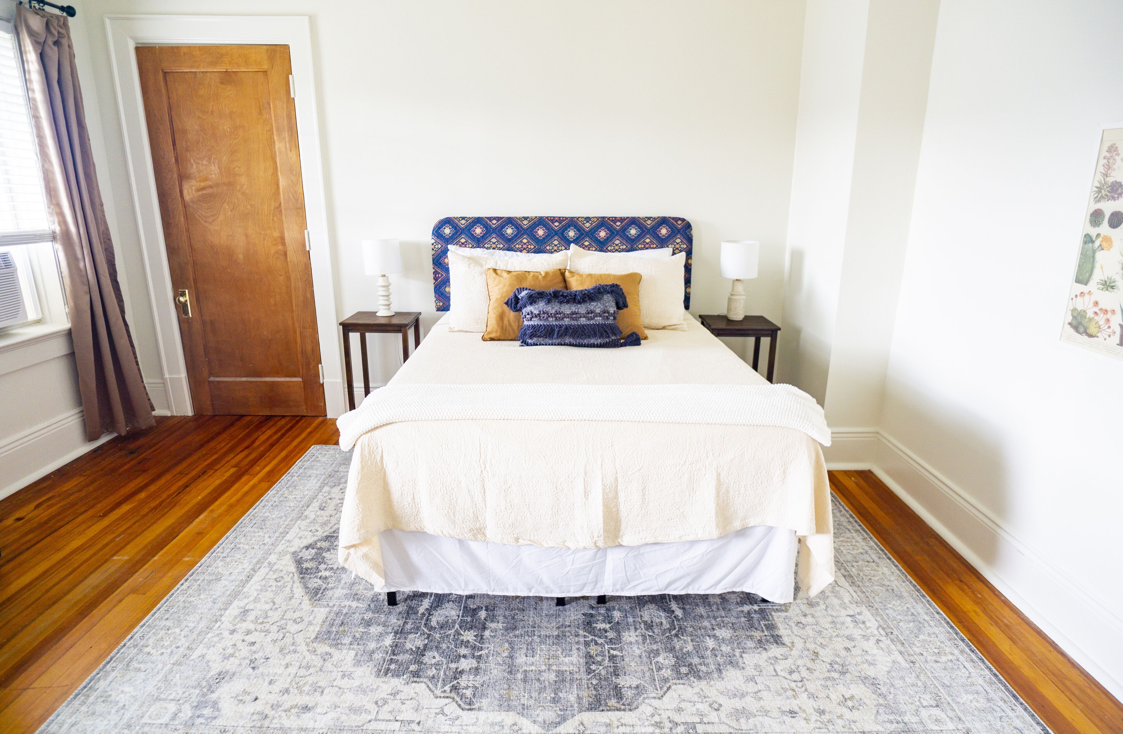 A neatly arranged bedroom with a wooden floor, a patterned rug, a bed with a blue headboard, and two bedside tables with lamps.