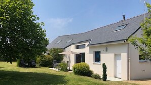 Une maison à deux étages avec un toit en pente, un petit jardin et un patio aménagé avec du mobilier d’extérieur.