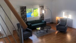 Un salon moderne avec un canapé vert, deux fauteuils et une table basse. Une lampe sur pied et une lampe placée sur une table d’appoint assurent l’éclairage. Une fenêtre offre une vue sur la verdure.