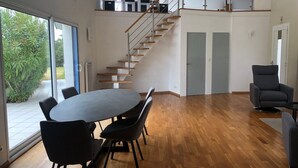 Un vaste espace de vie doté d’un plancher en bois, d’une table ronde avec des chaises, d’un escalier menant à un niveau supérieur et d’une grande porte-fenêtre coulissante donnant sur l’extérieur.